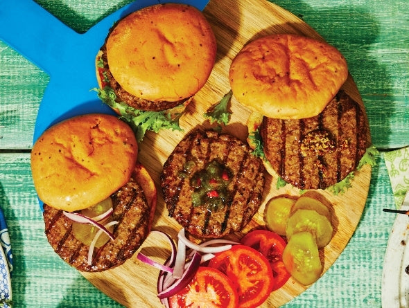 Trois burgers grillés sur une planche de bois avec légumes sur une surface verte