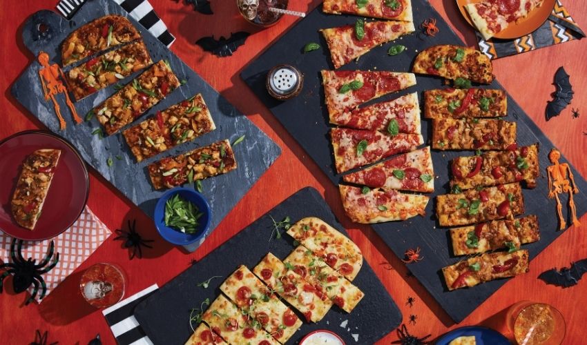 Four assorted flatbreads sliced into rectangular and square pieces, arranged on dark slate serving boards over a bright orange Halloween-themed table with spider decorations, black bats, and orange party accents.