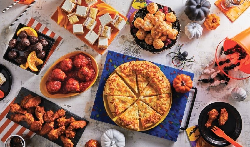 A styled Halloween party spread with a large sliced pizza in the center, surrounded by candy corn, jack-o'-lanterns, mini pumpkins, spider decorations, and assorted snacks on a white table.