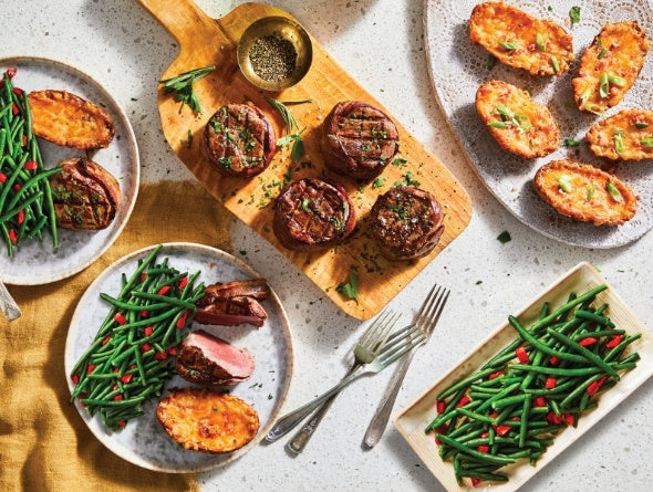 Table dressée avec assiettes de nourriture incluant haricots verts, pommes de terre rôties et biftecks sur une surface texturée