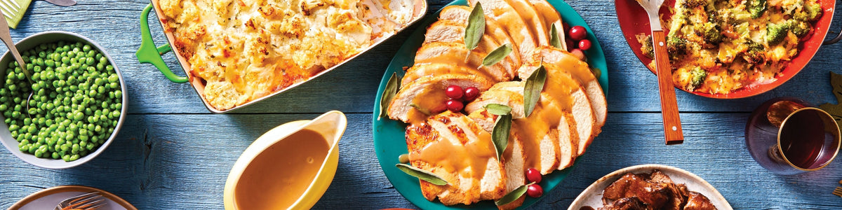 A top-down view of a Thanksgiving spread on a rustic blue wooden table, featuring sliced turkey with orange gravy, a casserole, green peas, stuffing, and a yellow gravy boat.
